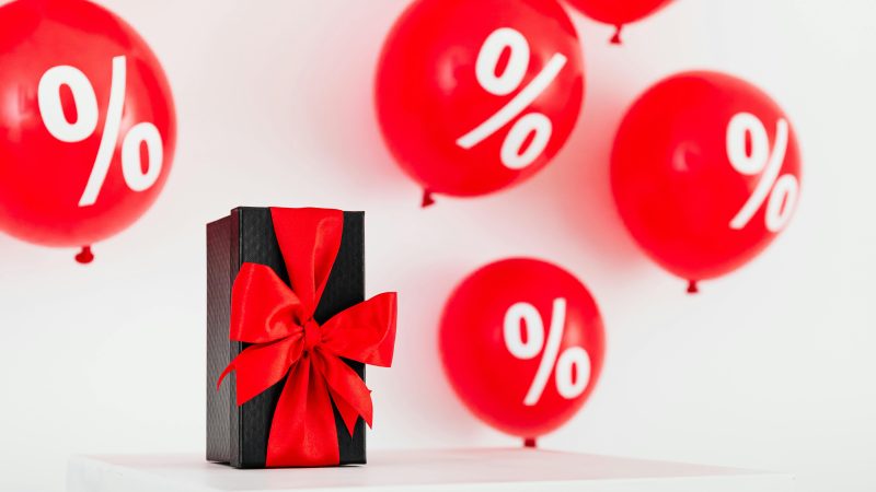 Stylish black gift box with red ribbon surrounded by red percentage balloons on a white background.